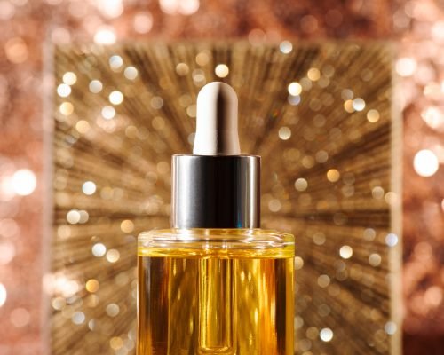 Face oil in glass bottle with silver cap and dropper on rose gold sequin background with starburst. Vertical front view. Self care and beauty for the christmas new year holiday season
